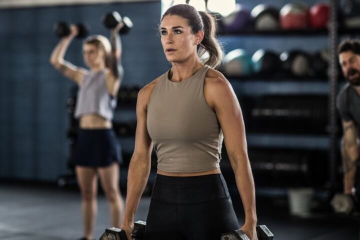 Woman stands holding dumbbells in a gym, people are working out behind her